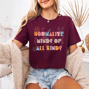 May include: A maroon t-shirt with the text "NORMALIZE MINDS OF ALL KINDS" in colorful, retro-style lettering. Floral and botanical illustrations surround the text. The shirt is short-sleeved.