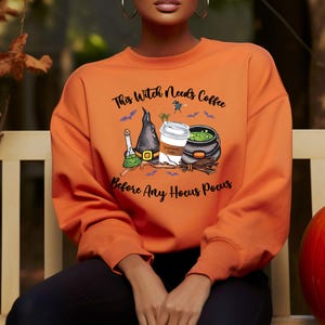 May include: Orange sweatshirt with a Halloween-themed graphic. The graphic includes a witch's hat, a coffee cup, a cauldron, and the text "This Witch Needs Coffee Before Any Hocus Pocus."