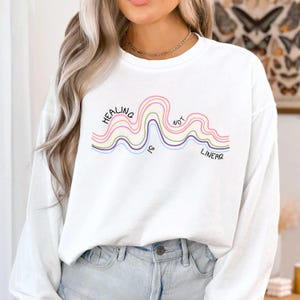 May include: A white long-sleeve sweatshirt featuring a graphic of wavy, multi-colored lines in pink, yellow, and purple. The text "HEALING IS NOT LINEAR" is printed in black, following the curves of the wavy lines across the chest area.
