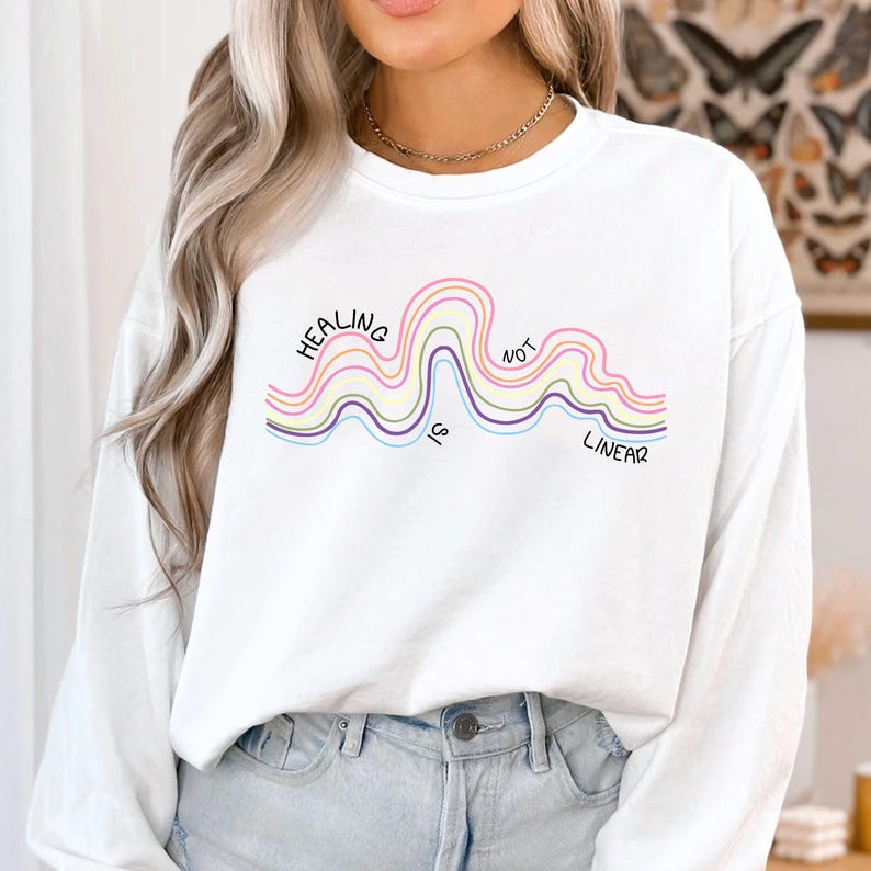 May include: A white long-sleeve sweatshirt featuring a graphic of wavy, multi-colored lines in pink, yellow, and purple. The text "HEALING IS NOT LINEAR" is printed in black, following the curves of the wavy lines across the chest area.