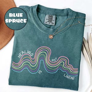 May include: A blue spruce colored t-shirt featuring a graphic of multi-colored wavy lines and the white text "HEALING IS NOT LINEAR". The shirt has a crew neckline and a Comfort Colors brand label on the inside collar indicating a size small.