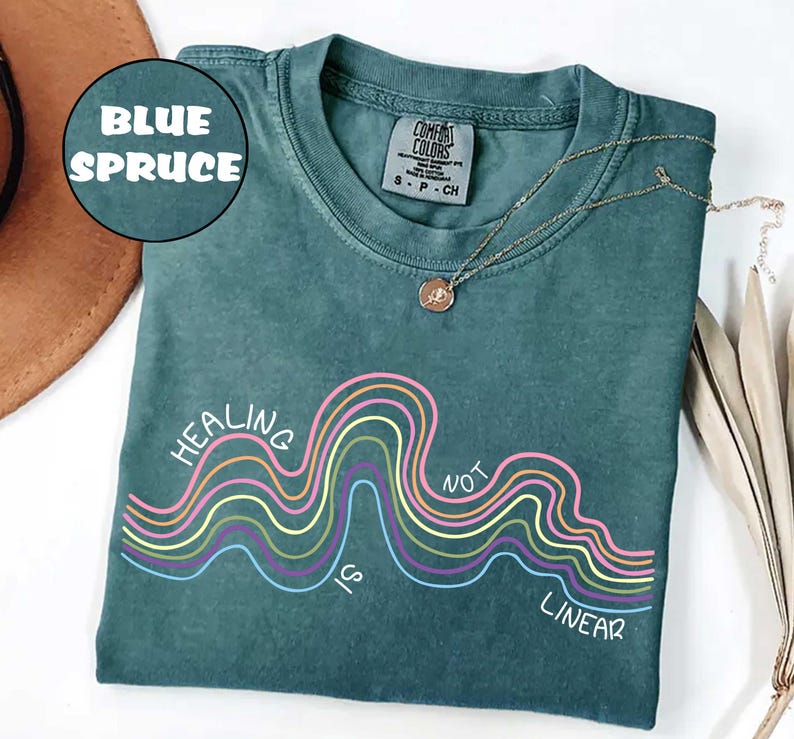 May include: A blue spruce colored t-shirt featuring a graphic of multi-colored wavy lines and the white text "HEALING IS NOT LINEAR". The shirt has a crew neckline and a Comfort Colors brand label on the inside collar indicating a size small.