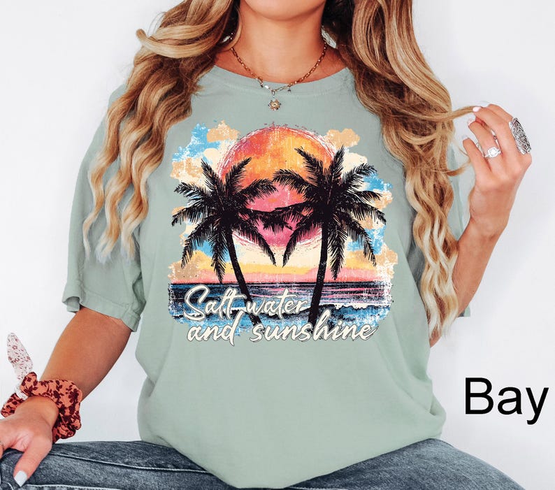 May include: A light green t-shirt featuring a colorful graphic of a sunset, palm trees, and ocean waves. The text "Salt water and sunshine" is printed below the image. The shirt is a relaxed fit.