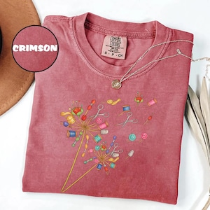 May include: Crimson-colored t-shirt with a dandelion design made of sewing notions. The design includes spools of thread, scissors, and buttons. A silver necklace with a pendant is draped across the shirt. The shirt has a Comfort Colors label.