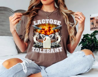 May include: Brown t-shirt with the words "LACTOSE TOLERANT" in a distressed font. The graphic features a milk jug, cows, fighter jets, and a fiery explosion. The shirt is a comfortable, casual style.