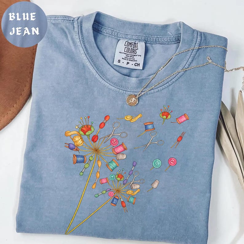 May include: A blue jean Comfort Colors t-shirt with a dandelion design made of sewing notions. The design includes spools of thread, scissors, and buttons. A silver necklace with a pendant rests on the shirt.