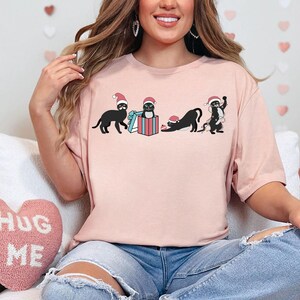 May include: A light pink t-shirt featuring a graphic of four black cats wearing Santa hats. One cat is standing, another is in a gift box, a third is stretching, and the last is tangled in Christmas lights. The shirt is worn by a person.