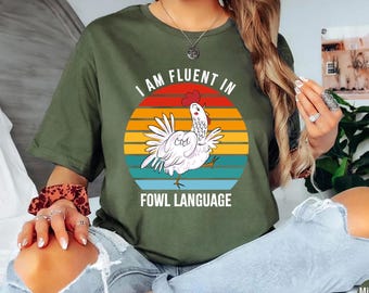 May include: An olive green t-shirt with the text "I AM FLUENT IN FOWL LANGUAGE" and a graphic of a white chicken in front of a sunset design. The shirt is made of a soft material.
