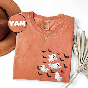 May include: Orange Comfort Colors t-shirt featuring a design of cartoon ghosts and bats. The shirt has a round neck and short sleeves. A gold necklace with a pendant is draped across the shirt. A brown hat is in the background.