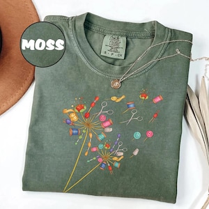 May include: Moss green t-shirt with a dandelion design made of sewing notions. The design includes spools of thread, scissors, and buttons in various colors. The shirt has a crew neck and short sleeves. The word "MOSS" is in a circle in the upper left corner.