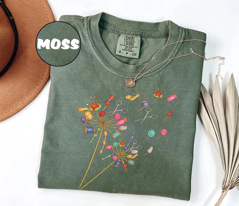 May include: Moss green t-shirt with a dandelion design made of sewing notions. The design includes spools of thread, scissors, and buttons in various colors. The shirt has a crew neck and short sleeves. The word "MOSS" is in a circle in the upper left corner.
