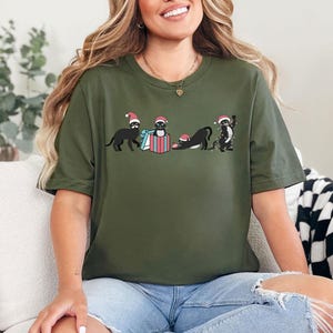 May include: Olive green t-shirt featuring a graphic of four black cats wearing Santa hats. One cat is standing, one is in a gift box, and two are in playful poses. The design includes a blue and red gift box. The shirt is a casual, festive apparel item.