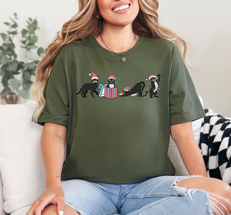 May include: Olive green t-shirt featuring a graphic of four black cats wearing Santa hats. One cat is standing, one is in a gift box, and two are in playful poses. The design includes a blue and red gift box. The shirt is a casual, festive apparel item.