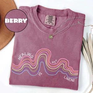 May include: A berry-colored short-sleeve t-shirt with a crew neckline. The shirt features a graphic of multi-colored wavy lines and the text "HEALING IS NOT LINEAR" printed in white. A silver-toned necklace rests on the collar of the shirt.