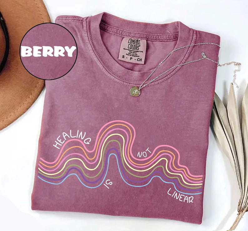 May include: A berry-colored short-sleeve t-shirt with a crew neckline. The shirt features a graphic of multi-colored wavy lines and the text "HEALING IS NOT LINEAR" printed in white. A silver-toned necklace rests on the collar of the shirt.