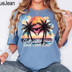 May include: A light blue t-shirt featuring a tropical sunset design with palm trees, a vibrant orange and yellow sun, and the text "Saltwater and sunshine". The shirt is made of a soft material.