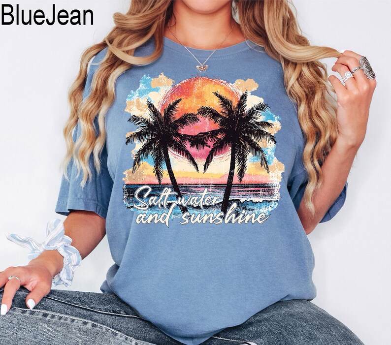 May include: A light blue t-shirt featuring a tropical sunset design with palm trees, a vibrant orange and yellow sun, and the text "Saltwater and sunshine". The shirt is made of a soft material.