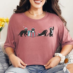 May include: A mauve-colored t-shirt featuring a graphic of four black cats wearing Santa hats. One cat stands, another sits in a gift box, and two others are in playful poses. The design is festive and whimsical.