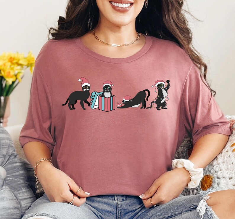 May include: A mauve-colored t-shirt featuring a graphic of four black cats wearing Santa hats. One cat stands, another sits in a gift box, and two others are in playful poses. The design is festive and whimsical.