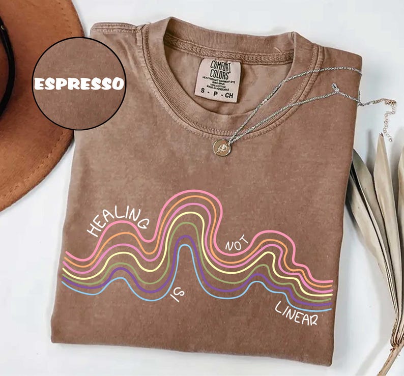 May include: A brown short-sleeved t-shirt with a crew neckline featuring a graphic of multi-colored wavy lines and the white text "HEALING IS NOT LINEAR". The shirt has a Comfort Colors brand label inside the collar indicating size small.