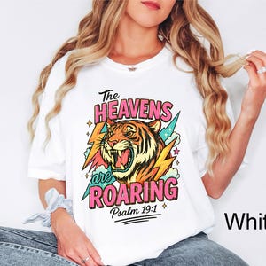May include: White t-shirt featuring a colorful graphic of a roaring tiger, lightning bolts, and the text "The Heavens are Roaring" with "Psalm 19:1". The design is in pink, orange, yellow, and teal.