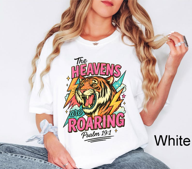 May include: White t-shirt featuring a colorful graphic of a roaring tiger, lightning bolts, and the text "The Heavens are Roaring" with "Psalm 19:1". The design is in pink, orange, yellow, and teal.
