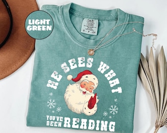 May include: Light green t-shirt with a Santa Claus graphic and the text "HE SEES WHAT YOU'VE BEEN READING". The Santa illustration features a red hat and gloves. A silver necklace with a pendant is also visible.