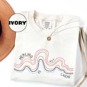 May include: An ivory t-shirt featuring a graphic of multi-colored wavy lines and the black text "HEALING IS NOT LINEAR". The shirt has a Comfort Colors brand label at the neck and is displayed with a gold-toned necklace featuring a small pendant.