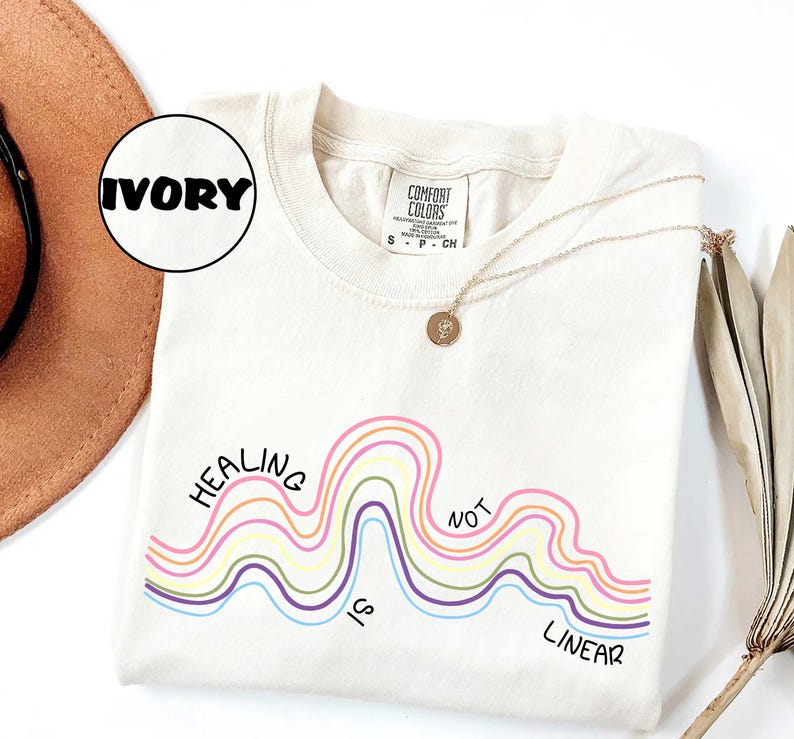May include: An ivory t-shirt featuring a graphic of multi-colored wavy lines and the black text "HEALING IS NOT LINEAR". The shirt has a Comfort Colors brand label at the neck and is displayed with a gold-toned necklace featuring a small pendant.