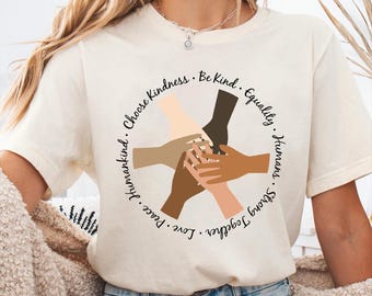 May include: A white t-shirt with a circle graphic of hands of different skin tones clasped together. The text around the circle reads "Strong Together. Love. Peace.  Humanity. Choose Kindness. Be Kind. Equality. Humans."