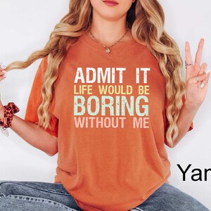Op de afbeelding: Een oranje t-shirt met de tekst "ADMIT IT LIFE WOULD BE BORING WITHOUT ME" in wit, groen en geel. Het shirt heeft een ronde hals en korte mouwen.
