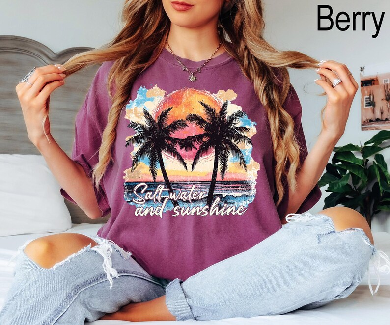 May include: Berry-colored t-shirt featuring a tropical sunset design with palm trees and the text "Salt-water and sunshine." The graphic is in shades of orange, yellow, blue, and black. The shirt is a relaxed fit.