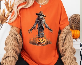 May include: Orange t-shirt featuring a Halloween-themed graphic. The design depicts a scarecrow figure with a skeletal face, wearing a hat and tattered clothing, standing atop a carved jack-o'-lantern. The background includes a fence and a spooky landscape.