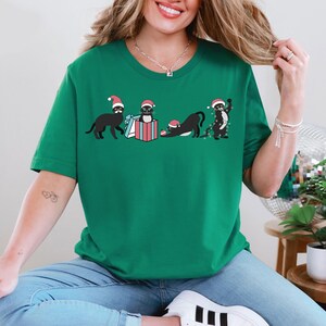 May include: Green t-shirt featuring a graphic of black cats wearing Santa hats. The cats are in various poses, including holding gifts and tangled in Christmas lights. The design is festive and playful, perfect for the holiday season.