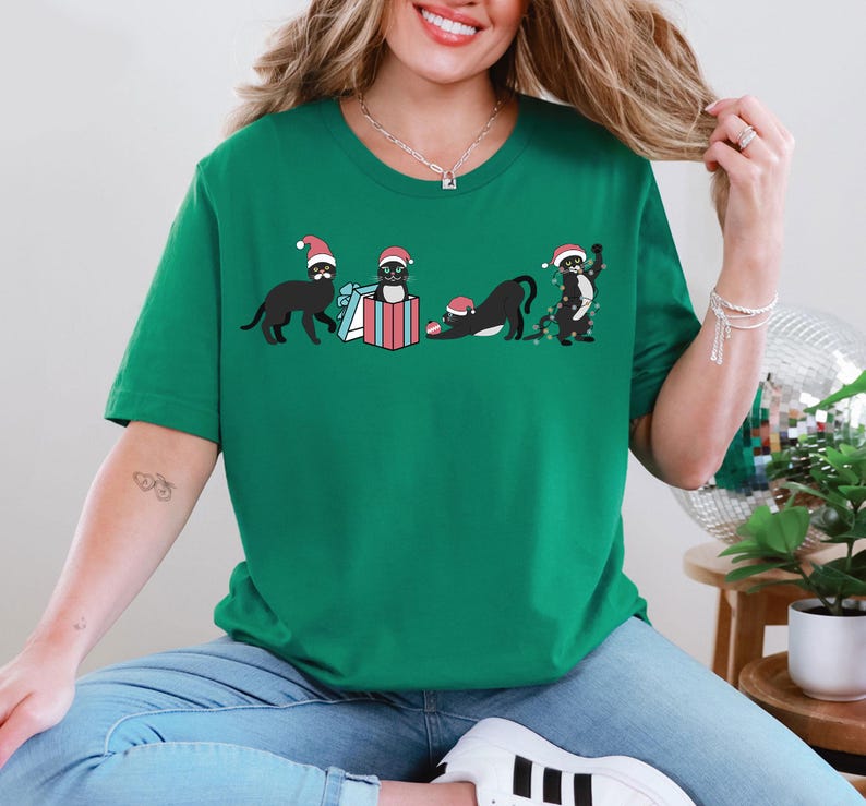 May include: Green t-shirt featuring a graphic of black cats wearing Santa hats. The cats are in various poses, including holding gifts and tangled in Christmas lights. The design is festive and playful, perfect for the holiday season.