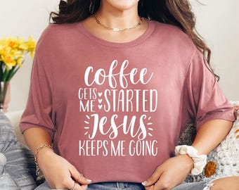 May include: A dusty rose-colored t-shirt with the white text "coffee gets me started Jesus keeps me going". The shirt has short sleeves and a round neckline. The text is in a handwritten style font.