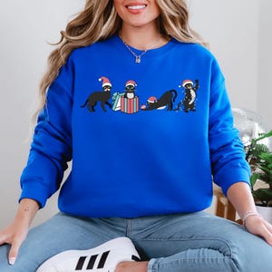 May include: Royal blue sweatshirt featuring a festive design of black cats wearing Santa hats. One cat carries a gift, another sits in a gift box, and a third stretches. The last cat is decorated with Christmas lights.