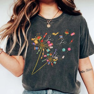 May include: Dark gray t-shirt featuring a colorful dandelion design made of sewing notions. The design includes spools of thread, scissors, buttons, and pins. The shirt has a vintage, worn look.