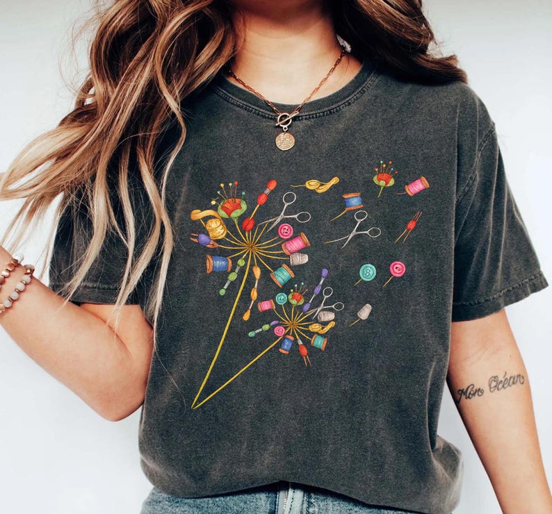 May include: Dark gray t-shirt featuring a colorful dandelion design made of sewing notions. The design includes spools of thread, scissors, buttons, and pins. The shirt has a vintage, worn look.