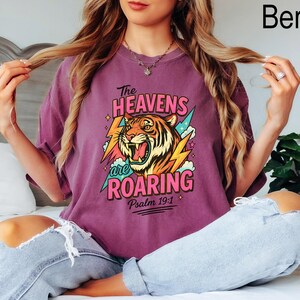 May include: Berry-colored t-shirt with a graphic of a roaring tiger, lightning bolts, and the text "The Heavens are Roaring" and "Psalm 19:1". The shirt is a relaxed fit.