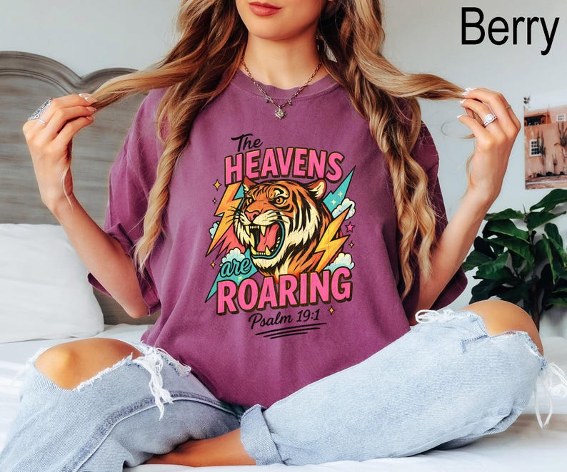 May include: Berry-colored t-shirt with a graphic of a roaring tiger, lightning bolts, and the text "The Heavens are Roaring" and "Psalm 19:1". The shirt is a relaxed fit.