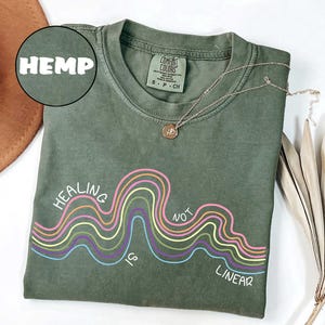 May include: An olive green crew neck t-shirt featuring a graphic of multi-colored wavy lines in pink, yellow, purple, and light blue. The text "HEALING IS NOT LINEAR" is printed in white around the wavy lines. A gold necklace rests on the collar.