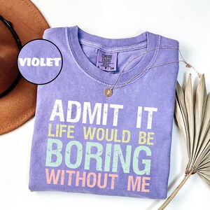 Op de afbeelding: Een violet Comfort Colors T-shirt met de tekst "ADMIT IT LIFE WOULD BE BORING WITHOUT ME" in wit, geel, groen en roze. Een gouden ketting en een bruine hoed staan ook op de afbeelding.
