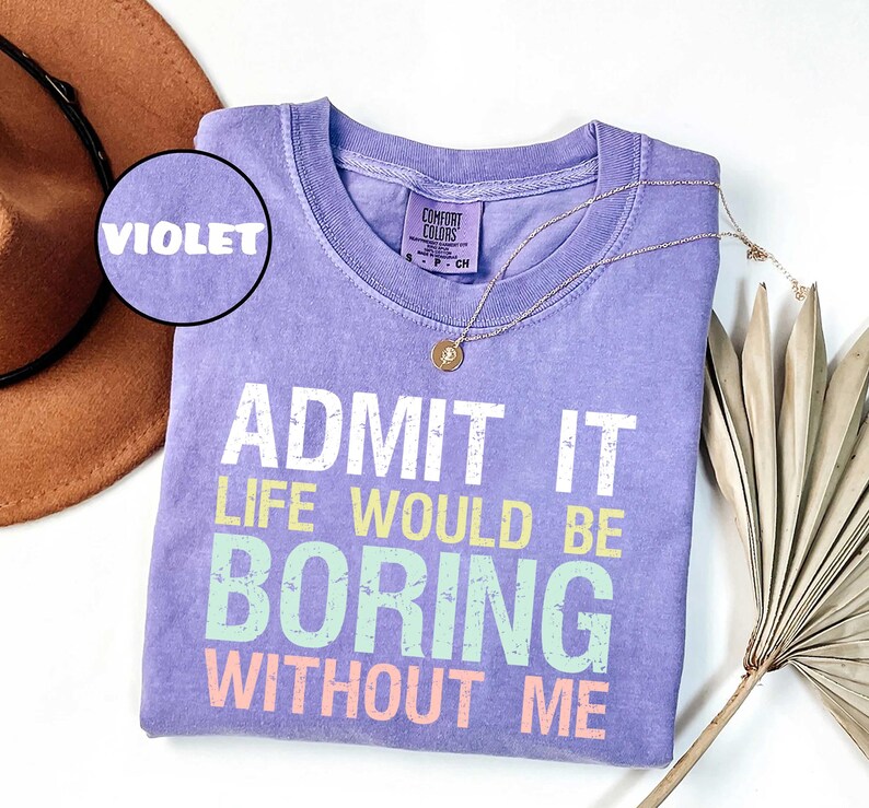 Op de afbeelding: Een violet Comfort Colors T-shirt met de tekst "ADMIT IT LIFE WOULD BE BORING WITHOUT ME" in wit, geel, groen en roze. Een gouden ketting en een bruine hoed staan ook op de afbeelding.