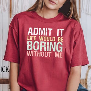 Op de afbeelding: Baksteenrood T-shirt met de tekst "ADMIT IT LIFE WOULD BE BORING WITHOUT ME" in wit, geel en groen. Het shirt heeft korte mouwen en een ronde hals.