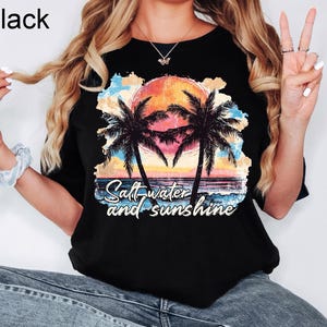 May include: Black t-shirt featuring a tropical sunset scene with palm trees and the text "Salt water and sunshine". The graphic includes a vibrant orange sun, blue water, and colorful clouds.