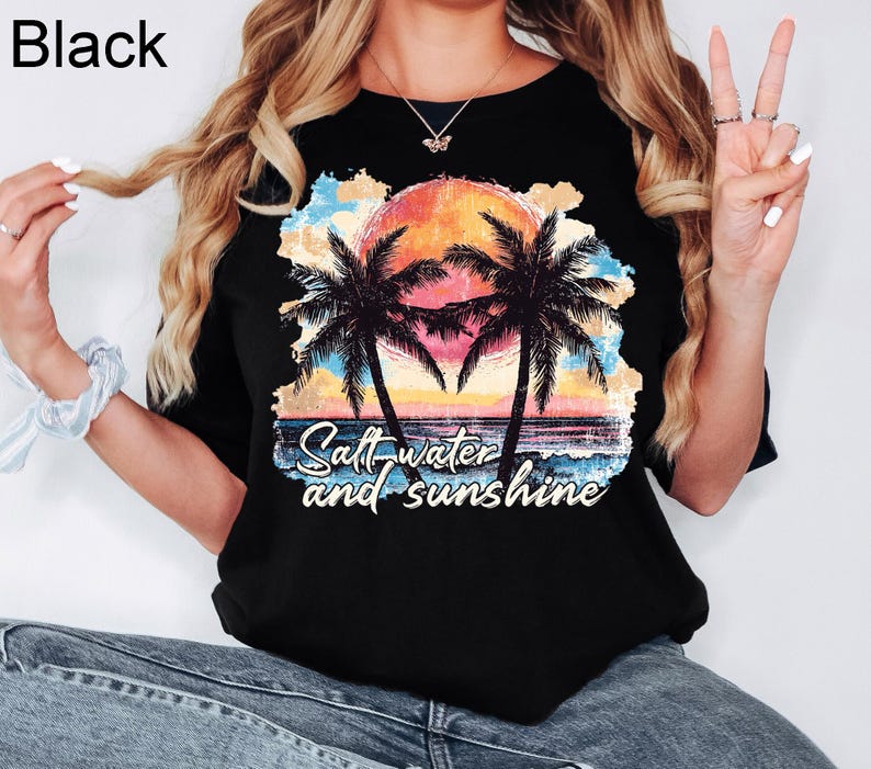 May include: Black t-shirt featuring a tropical sunset scene with palm trees and the text "Salt water and sunshine". The graphic includes a vibrant orange sun, blue water, and colorful clouds.