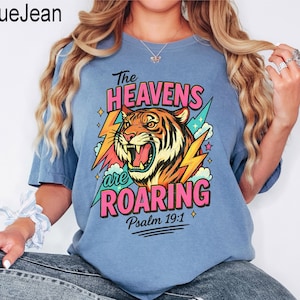 May include: A light blue t-shirt with a graphic of a roaring tiger, lightning bolts, and the text "The Heavens are Roaring" and "Psalm 19:1". The tiger is orange, black, and yellow. The text is pink, green, and white.