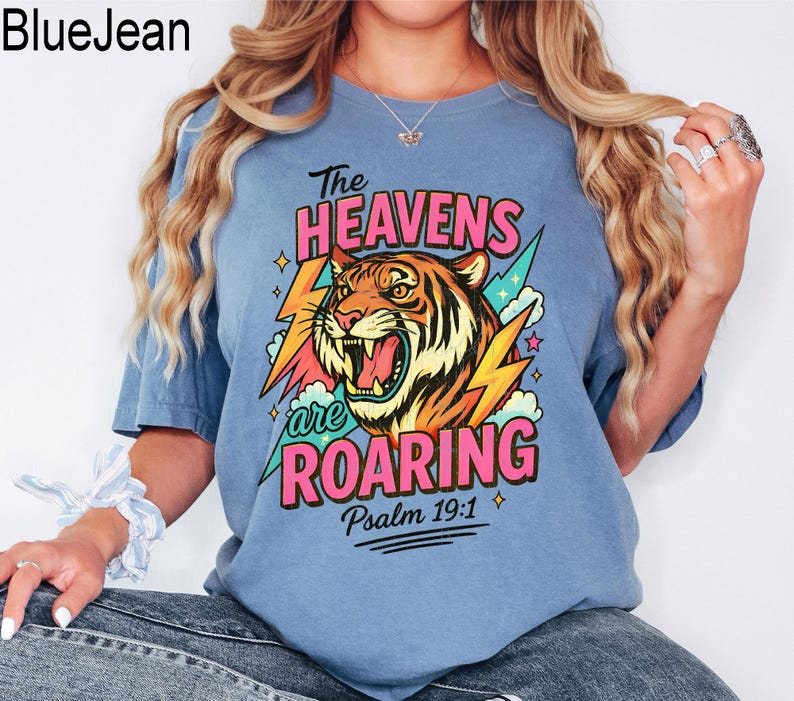 May include: A light blue t-shirt with a graphic of a roaring tiger, lightning bolts, and the text "The Heavens are Roaring" and "Psalm 19:1". The tiger is orange, black, and yellow. The text is pink, green, and white.