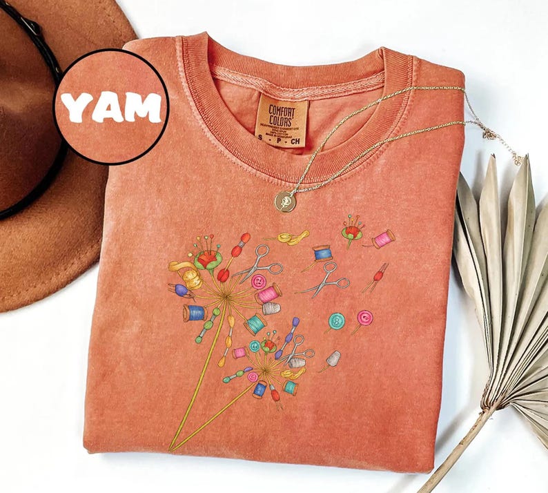 May include: A coral-colored Comfort Colors t-shirt featuring a dandelion design made of sewing notions. The design includes spools of thread, scissors, and buttons. A gold necklace with a pendant is draped on the shirt. The word "YAM" is in a circle in the upper left corner.